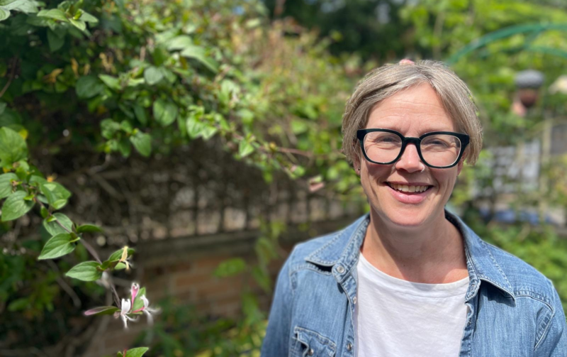 Professor Charlotte Ross wearing black framed glasses and a blue shirt is standing in the garden
