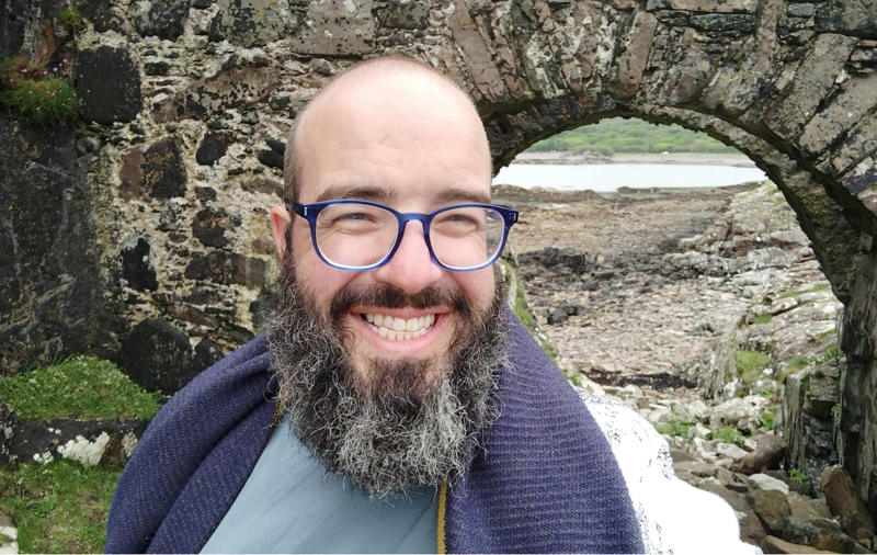 Jonny Tridgell wearing glasses and a blue top stands in front of an old grey stone bridge  