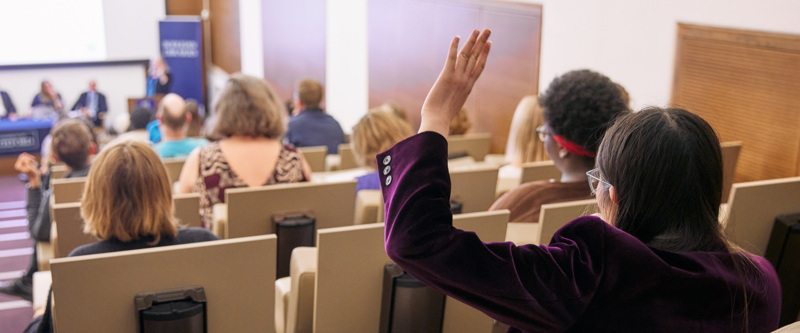 A student wearing a plum coloured jacket raising her hand in a lecture theatre