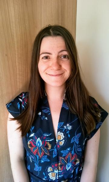 A photo of Jessica Boland, who is smiling at the camera. She has long brown hair and is wearing a navy top with multicoloured flowers