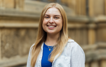 Lauren Schaefer, VP PG Education and Access, Oxford University Student’s Union Lauren Schaefer standing in front of college buildings in Radcliffe Square wearing a blue shirt