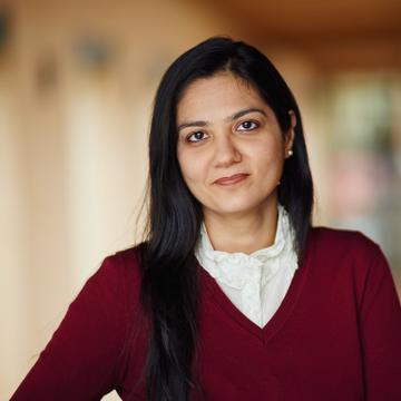 An image of Mahima wearing a red jumper and white shirt, looking at the camera against a blurred yellow background. 