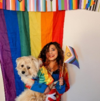A picture of charlie in front of a wall of pride flags, holding a dog wearing a rainbow jacket.