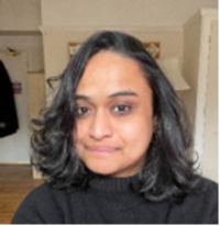 A picture of Sneha smiling at the camera. She is wearing a black turtleneck jumper.