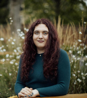 An image of Jessie sitting outside in nature, smiling at the camera. She has long red hair and is wearing a blue top. 