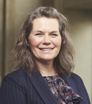 Katie is smiling at the camera against a neutral background. She has shoulder-length brown hair, and is wearing a dark blazer and a floral shirt. 