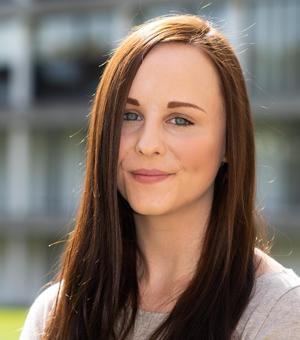 A photo of Clare smiling at the camera against an outdoor background. Clare has long brown hair, blue eyes and is wearing a grey top. 