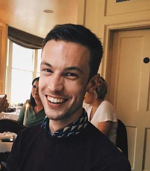 An image of James Ross smiling in a restaurant, wearing a jumper and a patterned shirt. 