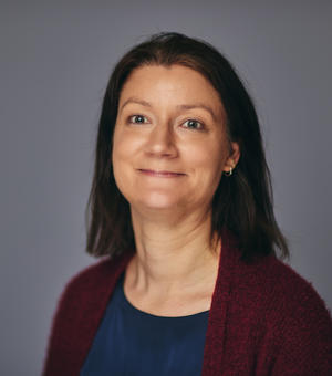 Jude smiling at the camera, against a grey background. She has medium length brown hair, is wearing a blue shirt and a red cardigan. 