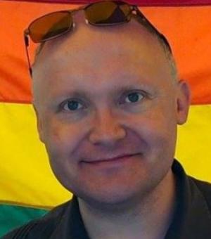 An image of Tony Brett in front of a Pride Flag. 