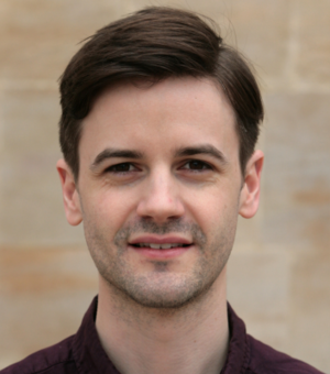 An image of Nathan smiling at the camera, against a brick wall. He has brown hair and is wearing a brown shirt. 