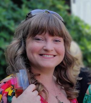 Scarlett is smiling at the camera. In the background is greenery. She has shoulder-length brown hair, and is wearing a floral top and sunglasses. She is holding a glass of wine. 