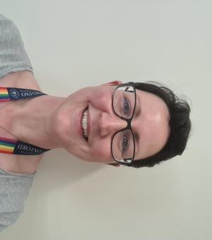 Rachel Lloyd in front of a blank wall, smiling at the camera and wearing a rainbow lanyard